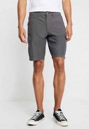Mann trägt graue Cargo-Shorts, weißes Hemd und schwarze flache Sneaker, steht vor einem weißen Hintergrund.
