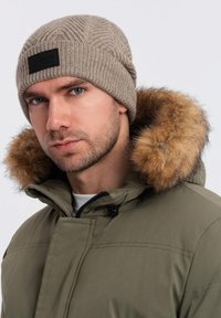 Gorro de punto beige con un diseño texturizado, que presenta una etiqueta negra. Se lleva con una chaqueta verde oliva que tiene una capucha forrada de piel.