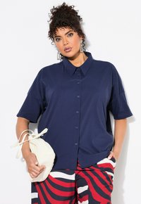 Maglietta blu navy a maniche corte con chiusura a bottoni, vestibilità comoda, abbinata a pantaloni a fantasia rossa, bianca e nera e una borsa color crema.