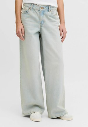 Světle modré džíny s širokými nohavicemi, nošené s bílými teniskami a bílým topem, zobrazené od pasu po nohy na jednobarevném pozadí.