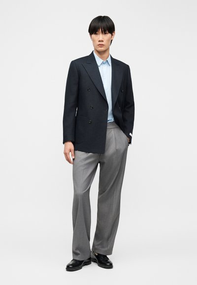 Άνδρας που φοράει σκούρο μπλε σακάκι διπλής κουμπιάς, ανοιχτό μπλε πουκάμισο, γκρι παντελόνι με πτυχώσεις και μαύρα δερμάτινα παπούτσια, στέκεται με το ένα χέρι στην τσέπη.