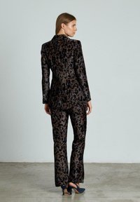 Abito con stampa leopardata realizzato in tessuto lucido; presenta un blazer aderente e pantaloni dritti, nelle tonalità di marrone e nero con dettagli in bottoni.