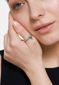 Anello d'argento a forma di cuore con una finitura liscia. Indossato sulla mano, mette in mostra un design delicato e una superficie lucida.