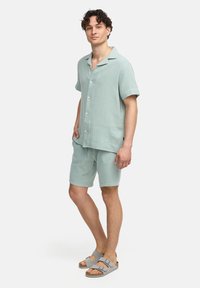 Completo in tessuto con camicia e pantaloni corti di colore verde chiaro, camicia a maniche corte con bottoni, abbinato a sandali grigi con doppia fibbia.