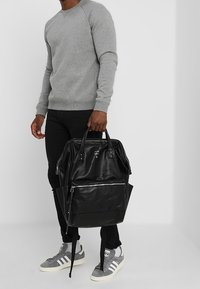 Mochila de cuero negro con dos asas superiores, bolsillo frontal con cremallera y correas ajustables, combinada con sudadera gris y pantalones negros.