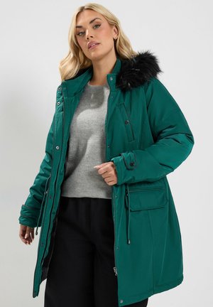 Femme portant un manteau parka vert avec capuche en fourrure noire, pull gris et pantalon noir sur un fond clair uni.