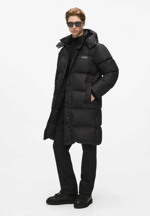 Man draagt een lange zwarte donsjas met capuchon, zwarte broek, zwarte schoenen en donkere zonnebril, staat met één hand in de zak.