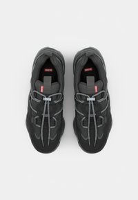 Black sneakers with gray accents and flexible laces, featuring a red logo on the tongue, displayed on a light background.