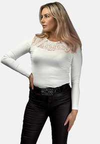Biała bluzka z długim rękawem z koronkowym karczkiem, w zestawieniu z dopasowanymi czarnymi spodniami. Zauważalne detale to czarny zdobiony pasek oraz gładka faktura.