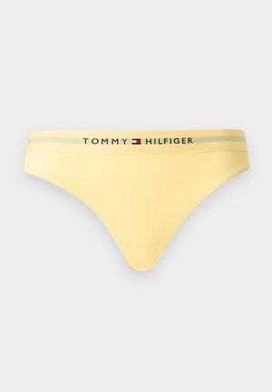 Žlté bikiny nohavičky s nízkym pásom a elastickým pásom s logom Tommy Hilfiger a vlajkou v čiernej, bielej, červenej a modrej farbe.