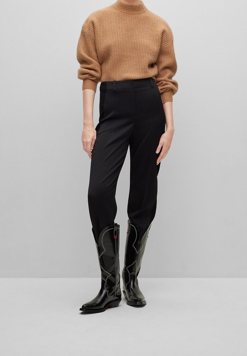 Pull en maille marron, pantalon noir ajusté et bottes de cowboy noires avec des surpiqûres blanches et une finition brillante.
