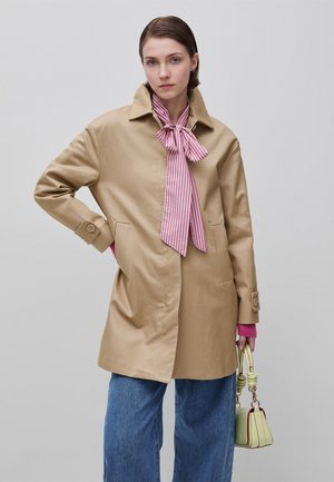 Donna con cappotto beige e cravatta rosa a strisce, maniche rosa visibili, jeans blu, che tiene una piccola borsa verde chiaro su sfondo semplice.