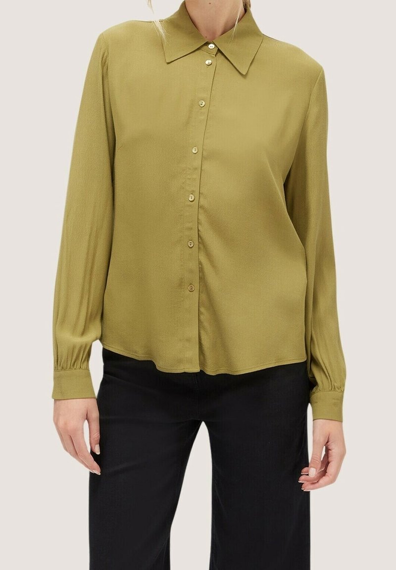 Femme portant un chemisier boutonné vert olive à manches longues avec un col pointu et un pantalon noir, vue du cou aux hanches.