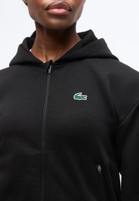 Κοντινό πλάνο ατόμου που φοράει μαύρο φούτερ με φερμουάρ και πράσινο λογότυπο κροκόδειλου στο στήθος και τσέπη με φερμουάρ στο πλάι.