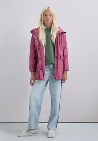 Parka impermeable rosa con capucha, botones negros, dos bolsillos frontales, superpuesta a una sudadera verde, combinada con jeans azul claros y zapatillas blancas.