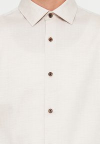 Light beige collared shirt with a subtle herringbone pattern and four visible brown buttons down the front.
