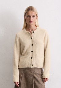 Blondynka w beżowym, teksturowanym swetrze zapinanym na guziki i brązowej spódnicy w kratę, stojąca przy gładkiej białej ścianie.