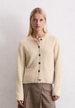 Femme blonde portant un cardigan boutonné en beige texturé et une jupe en tartan marron, se tenant devant un mur blanc uni.