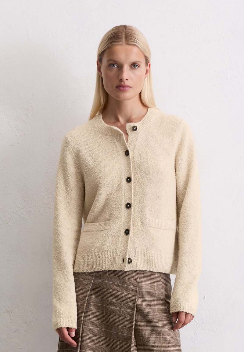 Blondynka w beżowym, teksturowanym swetrze zapinanym na guziki i brązowej spódnicy w kratę, stojąca przy gładkiej białej ścianie.