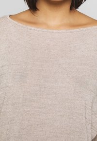 Beige neulepusero, jossa on leveä venepääntie, pehmeä rakenne ja väljä istuvuus. Sisältää hienovaraisia vaakaraitoja ja vähäiset saumat.