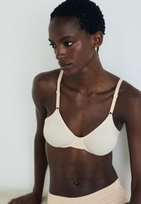 Nude krāsas krūšturis ar gludu audumu, trīsstūra cups un regulējamām siksnām, kuru rotā mazi metāla akcenti; minimālisma stils.