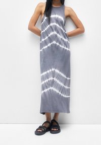 Šedé maxi šaty s tie-dye vzorem a bílými motivy, bez rukávů, vyrobené z měkké tkaniny, v kombinaci s tmavými pletenými sandály.