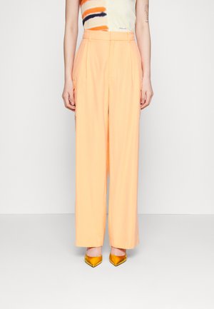 Pantalons taille haute de couleur pêche avec des jambes larges, présentant des plis sur le devant et des poches latérales, associés à des talons aiguilles jaunes.