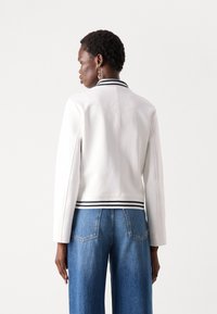 Veste bomber en cuir blanc avec un col et des poignets à rayures noires et blanches. Possède un design ajusté et une texture lisse.