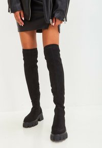 Zwarte boven-de-knie laarzen van zacht suède, met een chunky zool, een gladde textuur en een ritssluiting aan de zijkant.