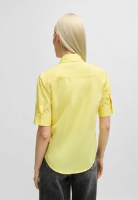 Kurzärmliges gelbes Hemd aus leichtem Stoff mit glatter Textur. Verfügt über einen klassischen Kragen und aufgerollte Ärmel, abgerundeten Rückenabschluss.