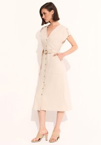 Beżowa lniana sukienka z krótkimi rękawami, dekoltem w serek i paskiem w talii. Posiada guziki z przodu oraz boczne kieszenie. Noszona z beżowymi sandałami na obcasie.