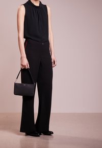 Person med ärmlös svart topp och svarta vida byxor med röd sidostrip, håller en liten svart handväska, står på beige golv.