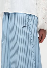 Άτομο που φορά φαρδιά μπλε και λευκή κάθετα ριγωτή παντελόνα με κεντημένο λογότυπο στον μηρό και λευκό μακρυμάνικο τοπ με δαντέλα με οπές.