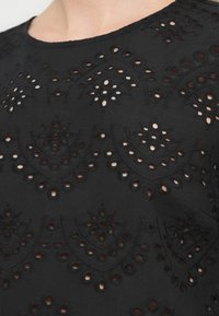 Μαύρο τοπ με περίτεχνες κεντητές περικοπές σε λουλουδένια μοτίβα, στρογγυλό λαιμόκοψη και υφασμάτινη υφή που δημιουργεί μία στρωματοποιημένη εμφάνιση.