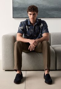 Marineblaues Hemd mit kurzen Ärmeln und Blumenmuster, beige Leinenhose, dunkle Loafers; sitzt auf einem hellgrauen Sofa mit strukturierter Polsterung.