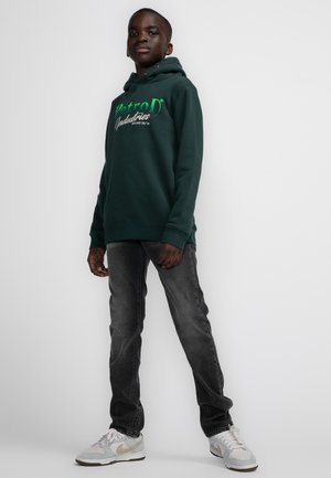 Donkergroene hoodie met voorlogo en trekkoordcapuchon, gecombineerd met vervaagde zwarte jeans en lichtgrijze sneakers met gouden accenten.