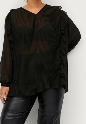 Kobieta nosząca przezroczystą czarną plisowaną bluzkę z falbankami po bokach i długimi rękawami, w połączeniu z czarnymi skórzanymi spodniami.