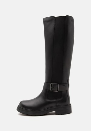 Bota negra de cuero hasta la rodilla con una correa con hebilla alrededor del tobillo y una suela gruesa de goma.