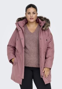 Pink parka jacket with a faux fur hood, zip and snap closures, and side pockets. Underneath, a fitted mauve sweater and black pants.