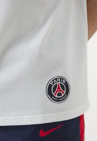 Camiseta de algodón blanca con un logo negro redondo que presenta "PARIS SAINT-GERMAIN" y un diseño de la Torre Eiffel en la parte inferior izquierda.
