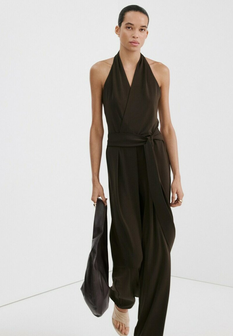 Frau trägt einen schwarzen, ärmellosen Halter-Overall mit weitem Bein, hält eine schwarze Tasche und steht vor weißem Hintergrund.