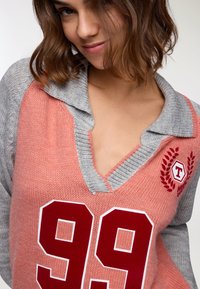 Maglione lavorato a maglia in corallo rosa e grigio, con scollo a V, colletto grigio e grande stampa rossa "99" con logo ricamato e accenti di alloro.