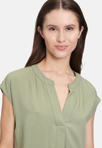 Kurzärmlige, olivgrüne Bluse mit V-Ausschnitt, weichem Material und dezenten Stichdetails an der Vorderseite und den Schultern.