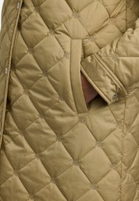 Padded, alyvuogių žalios spalvos striukė su deimantiniais raštais, siuvinėtomis logotipo detalėmis ir šoniniu kišeniu su mygtuko užsegimu.