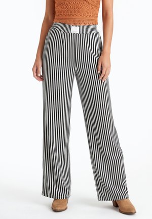 Vrouw draagt hooggetailleerde, wijdere zwarte en witte verticale gestreepte broek, bruine schoenen met hak en een kort oranje kanten top.