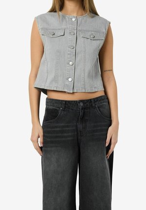 Femme portant une veste en jean grise courte sans manches avec des boutons et un jean noir taille haute, ample, debout les mains le long du corps.