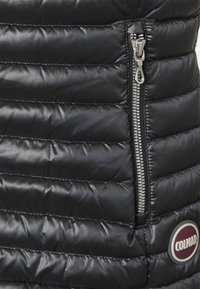 Black puffer vest with horizontal stitching, featuring a zippered side pocket and a circular red and white logo patch. Smooth, shiny texture.