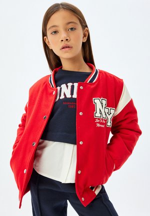 Rote Collegejacke mit weißen und marineblauen Akzenten, mit einem großen "NY"-Logo und Druckknöpfen. Getragen über einem marineblauen Sweatshirt.
