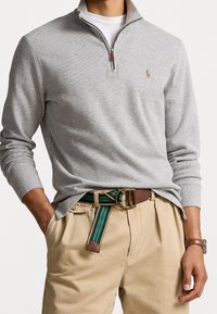 Šedý pletený svetr s límcem na čtvrtinový zip, s malým vyšitým logem. Kombinováno s béžovými kalhotami a páskem s mnoha pruhy.