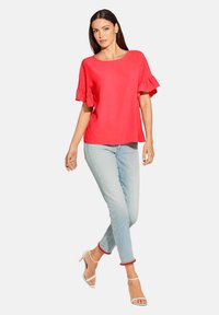 Rotes kurzärmeliges Oberteil mit Rüschensäumen, kombiniert mit hellblauen Jeans mit rotem Pompon-Besatz an den Säumen und weißen höhenverstellbaren Sandalen.